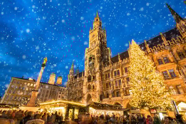 Belebter Weihnachtsmarkt vor einem historischen Rathaus mit grossem geschmücktem Weihnachtsbaum und fallendem Schnee.