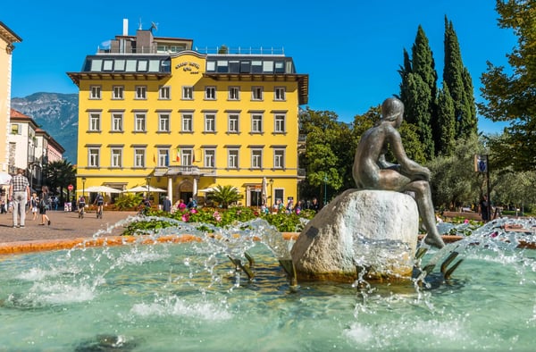 Brunnen mit Skulptur vor einem gelben Hotel in Riva del Garda bei sonnigem Wetter