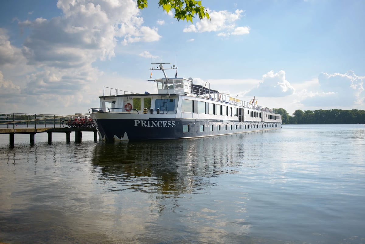 Flusskreuzfahrtschiff MS Princess liegt an einem Steg auf ruhigem Wasser, umgeben von Natur, unter blauem Himmel mit Wolken