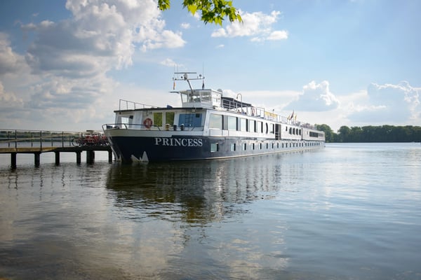 Flusskreuzfahrtschiff MS Princess liegt an einem Steg auf ruhigem Wasser, umgeben von Natur, unter blauem Himmel mit Wolken