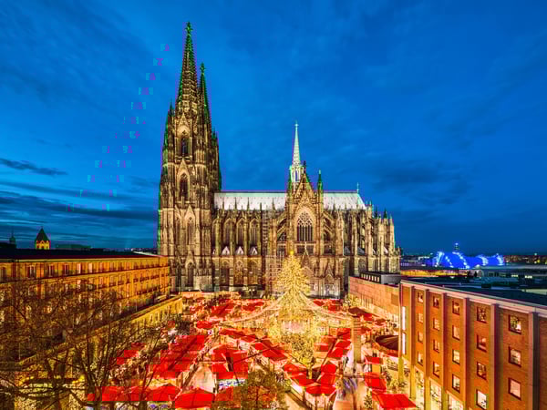 Weihnachtsmarkt vor dem Kölner Dom bei Abenddämmerung mit beleuchtetem Tannenbaum und roten Marktständen.