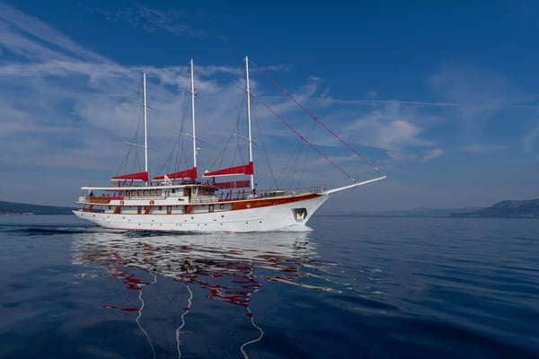 Navire de croisière à voiles blanc avec trois mâts et des voiles rouges naviguant par mer calme sur une eau bleue sous un ciel légèrement nuageux