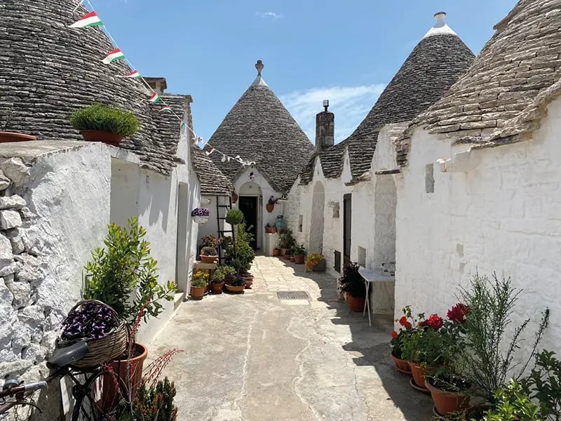 Une ruelle étroite à Alberobello bordée de trulli traditionnels aux murs blancs et aux toits de pierre coniques, décorée de pots de fleurs sous un ciel bleu