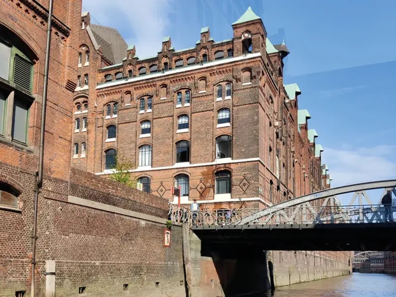 Historisches rotes Backsteingebäude an einem Kanal mit kleiner Brücke im Vordergrund und blauem Himmel