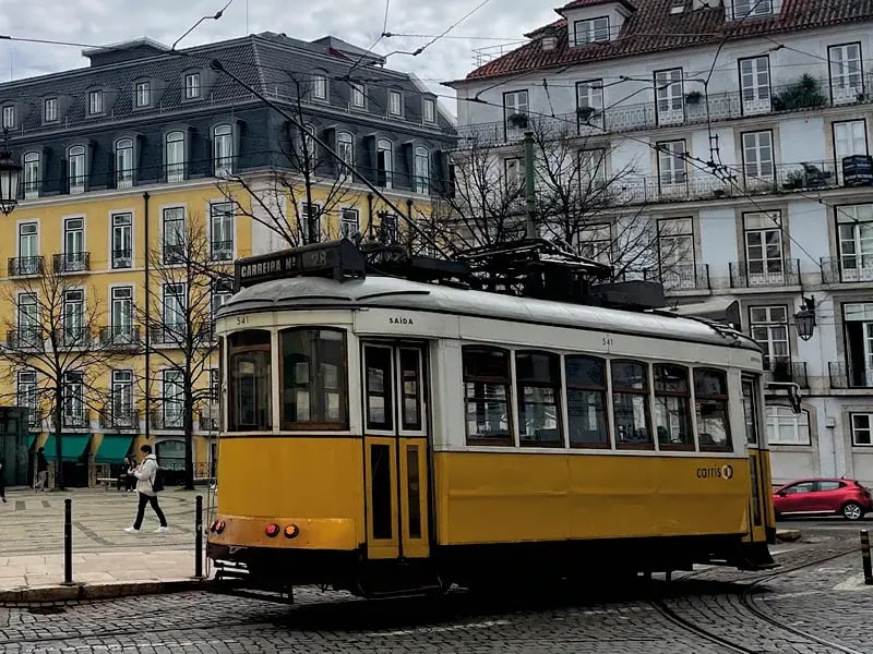 Eine klassische gelbe Straßenbahn der Linie 28 fährt über das Kopfsteinpflaster eines Platzes in Lissabon vor historischen Gebäuden