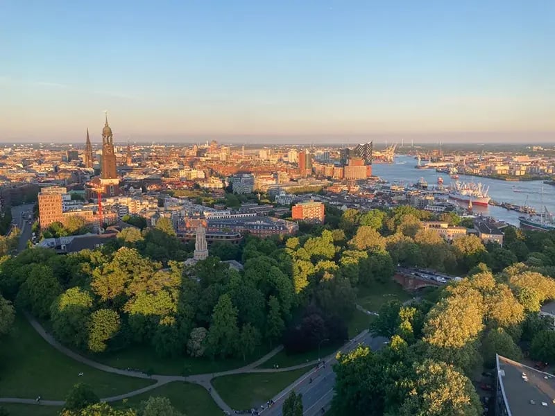 Stadtansicht Hamburg mit Park im Vordergrund, Hafen und Fluss sowie Hochhäusern im Hintergrund bei Abendlicht