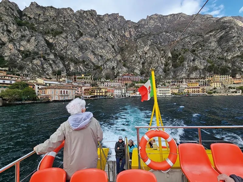Person steht am Heck eines Bootes auf einem See, im Hintergrund ein Ort am Ufer vor steilen Felsen