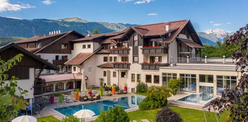 Aussenansicht eines modernen Alpenhotels mit mehreren Gebaeuden, Balkonen und Gartenanlage, davor ein Aussenpool mit Liegen, umgeben von Bergen und blauem Himmel.