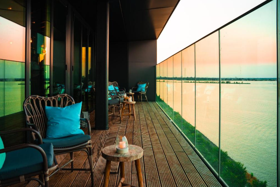 Moderne Skybar mit Bartresen und großer Fensterfront, dahinter eine Terrasse mit Stühlen und Blick auf das IJmeer.