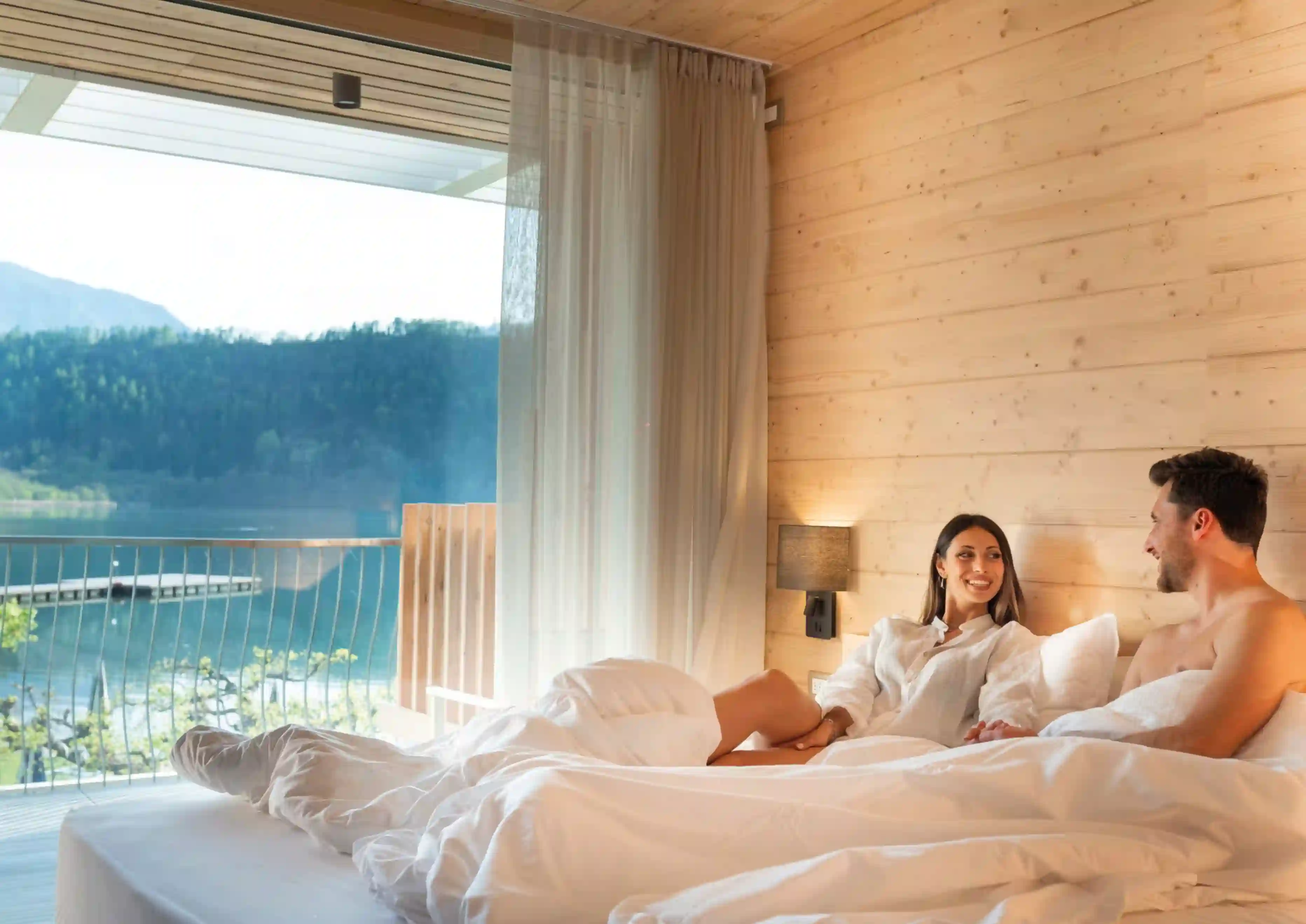 Paar entspannt in hellem Hotelzimmer mit Holzverkleidung, grossem Bett und Balkon mit Blick auf See und Berge.