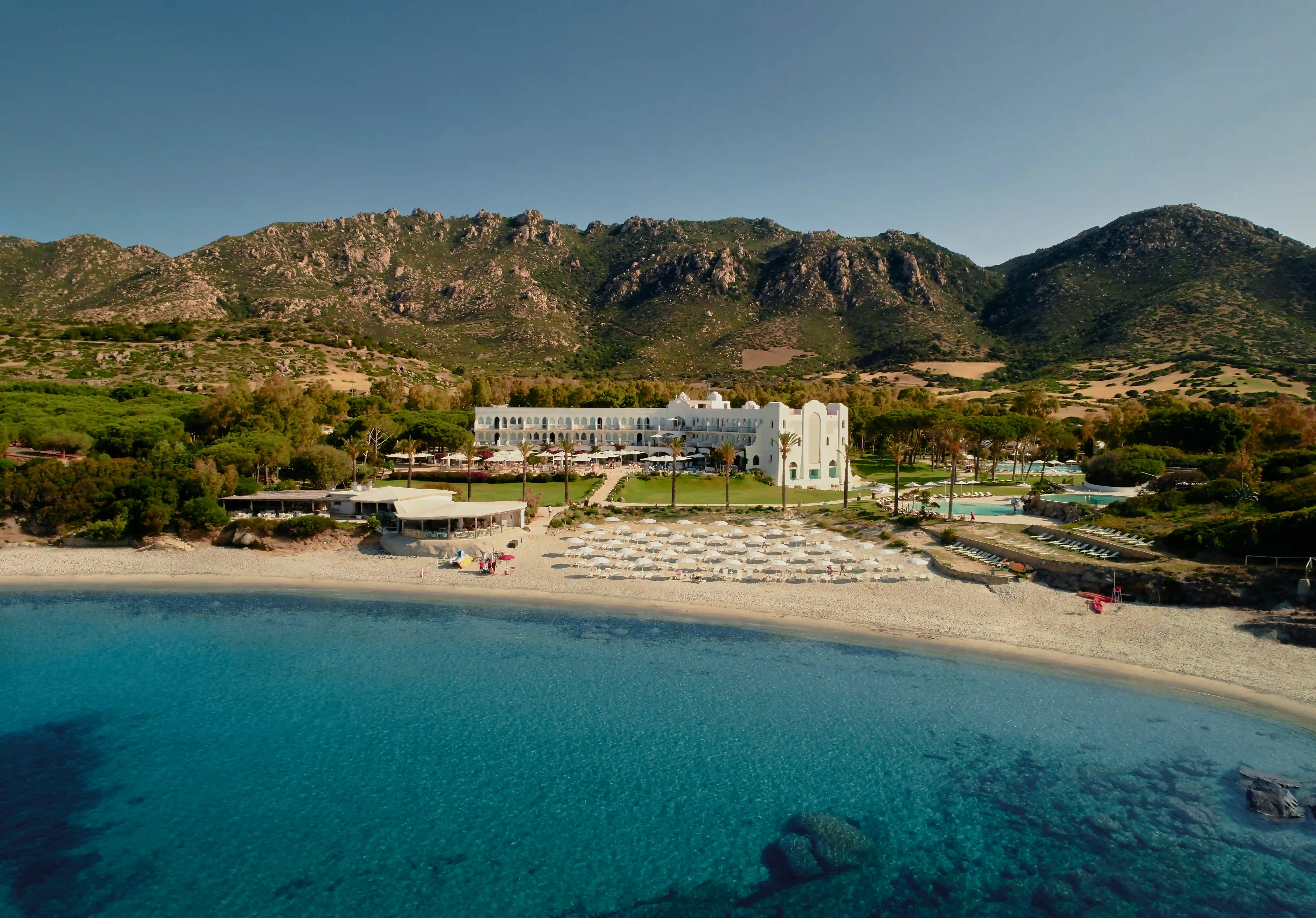 Panoramablick auf den Privatstrand des Hotels Falkensteiner Capo Boi auf Sardinien mit türkisfarbenem Meer, heller Sandbucht, geordneten Sonnenliegen und Palmen vor einer gruenen Kuestenlandschaft