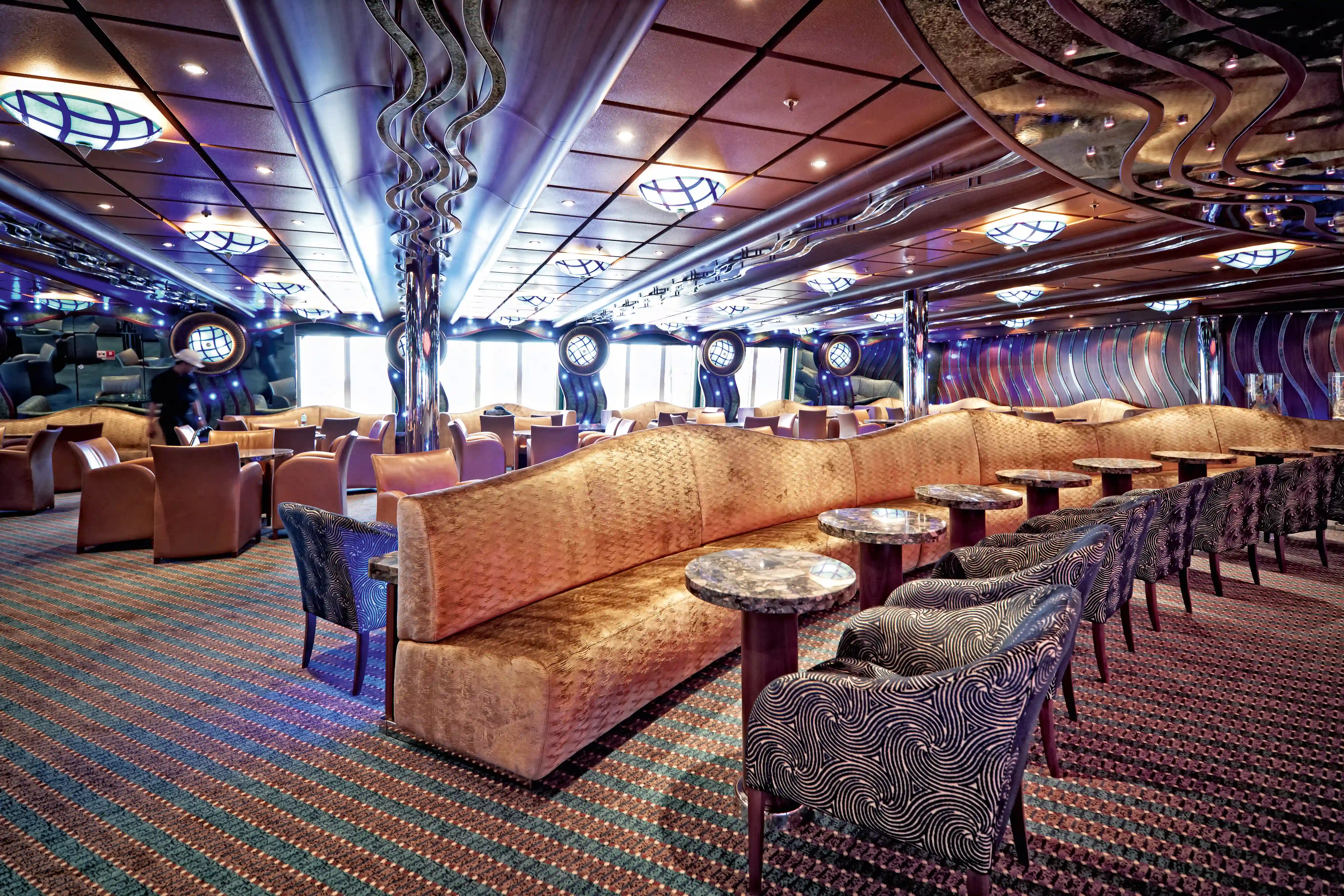 Grosszuegige Lounge mit gepolsterten Sitzbänken, kleinen Tischen und dekorativer Beleuchtung im Innenbereich des Schiffs