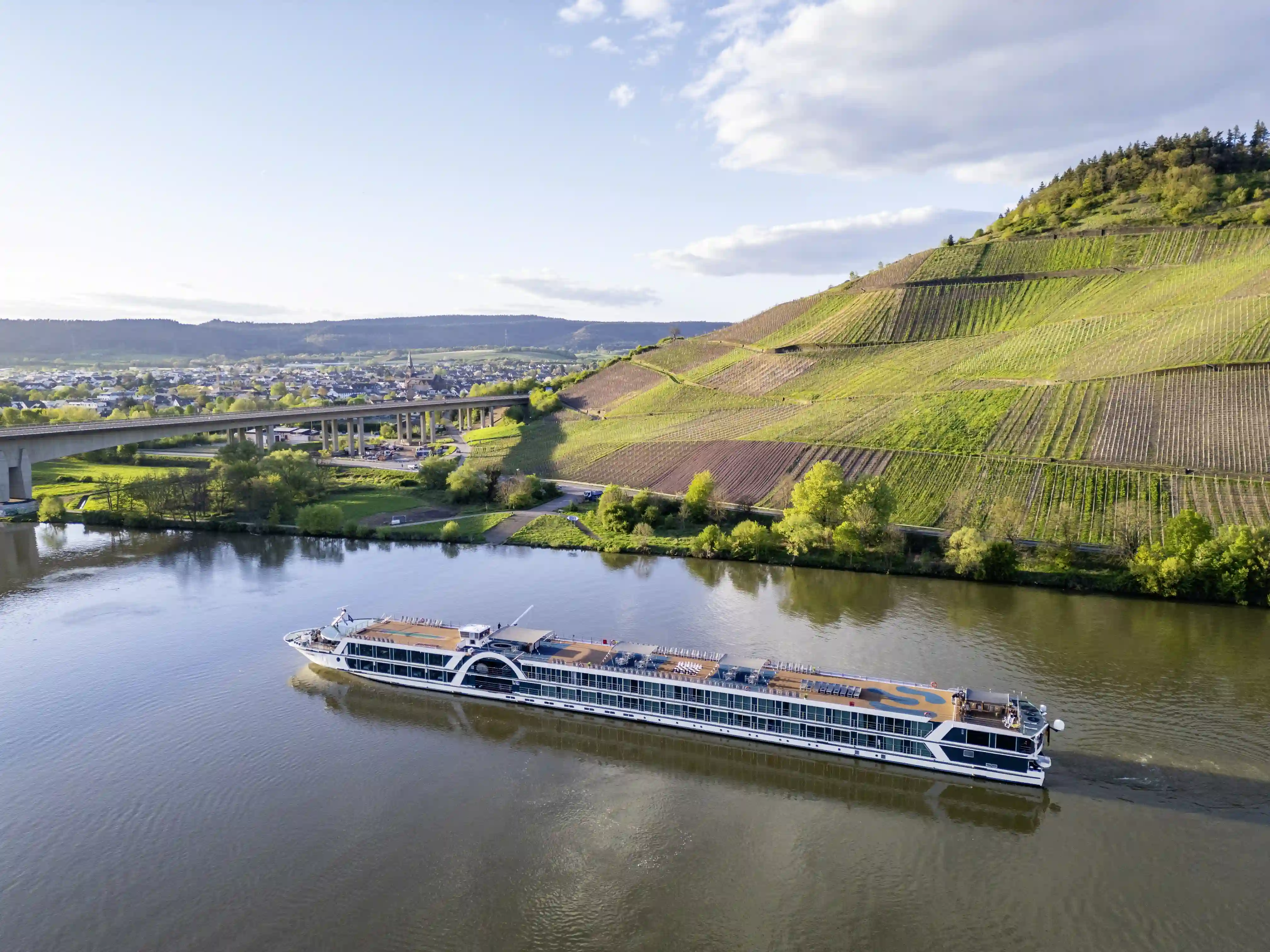 Luftaufnahme des Flusskreuzfahrtschiffs Elegant auf einem breiten Fluss mit Weinbergen, Brücke und Ortschaft im Hintergrund