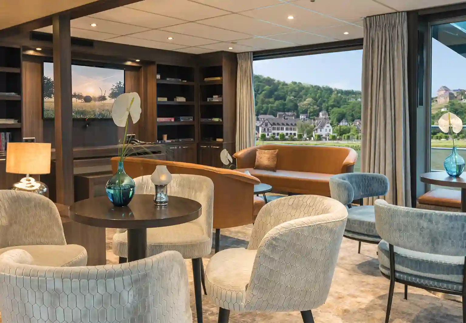 Eleganter Clubbereich mit Sesseln, Sofas, Regalen, warmem Licht und Blick auf eine Ortschaft am Flussufer
