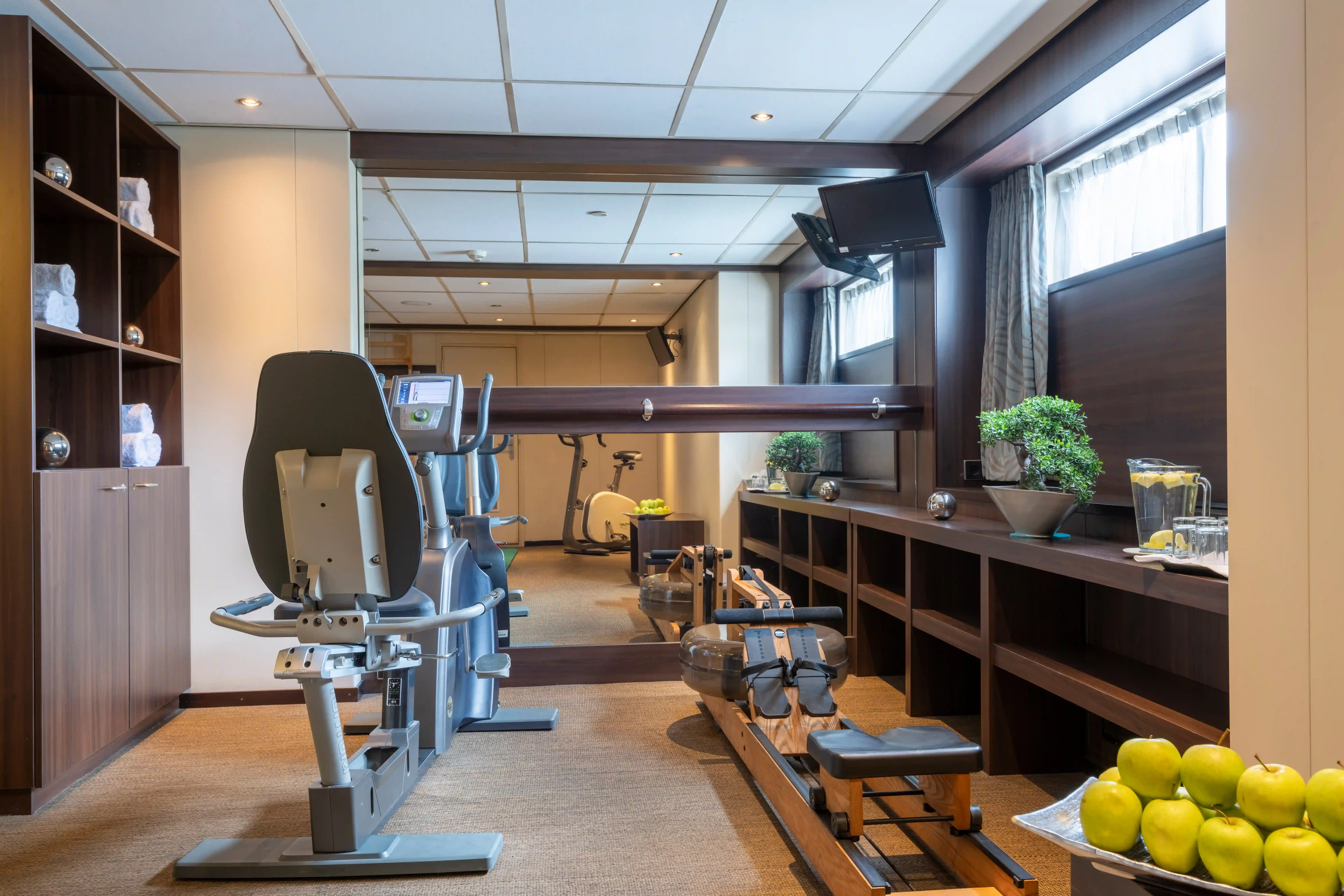 Fitnessraum mit Trainingsgeräten, Spiegelwand, Handtüchern, Wasser und Obst zur Erfrischung