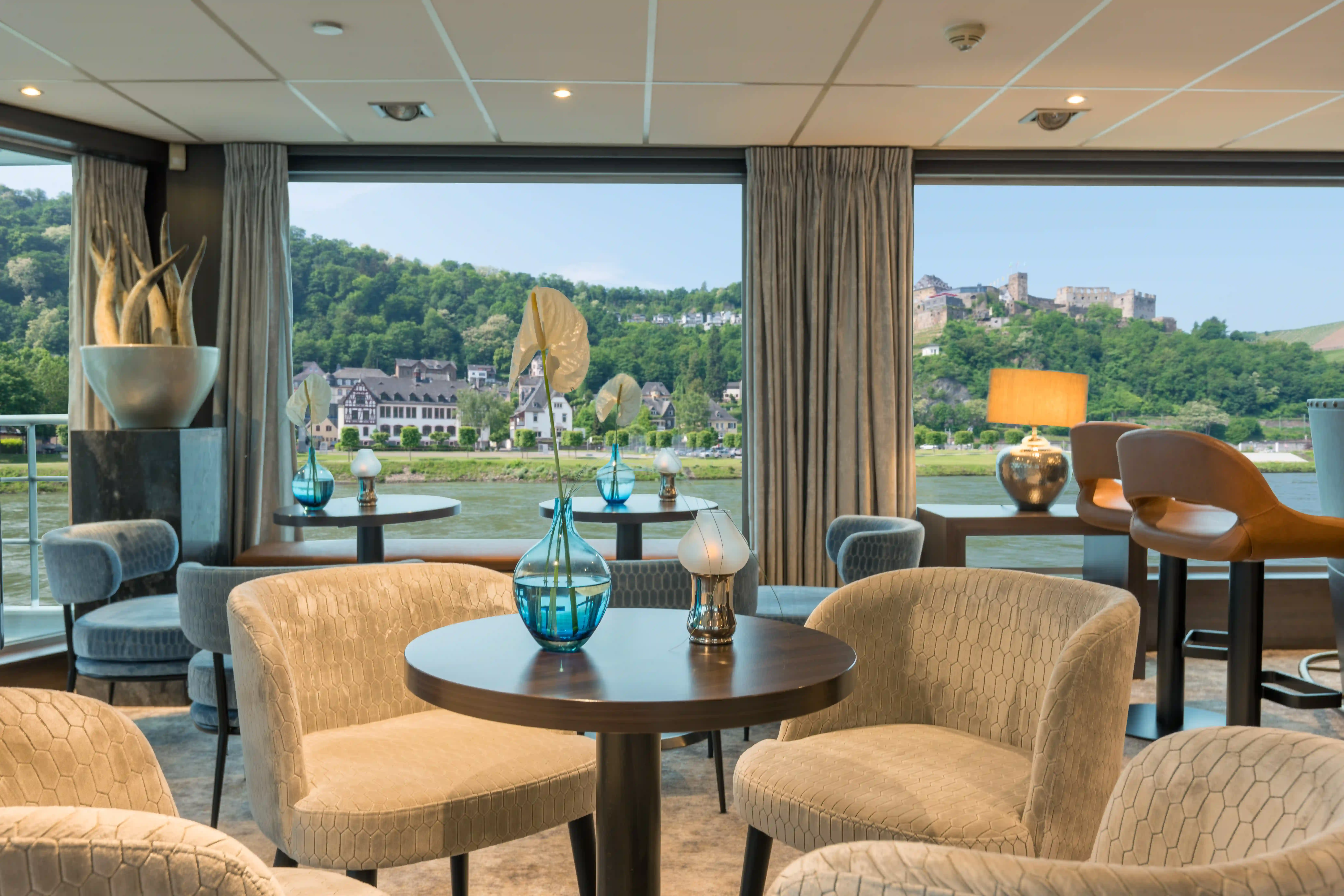 Panorama Bar mit runden Tischen, gepolsterten Sesseln, Vasen und weitem Blick auf Fluss und Burgenlandschaft