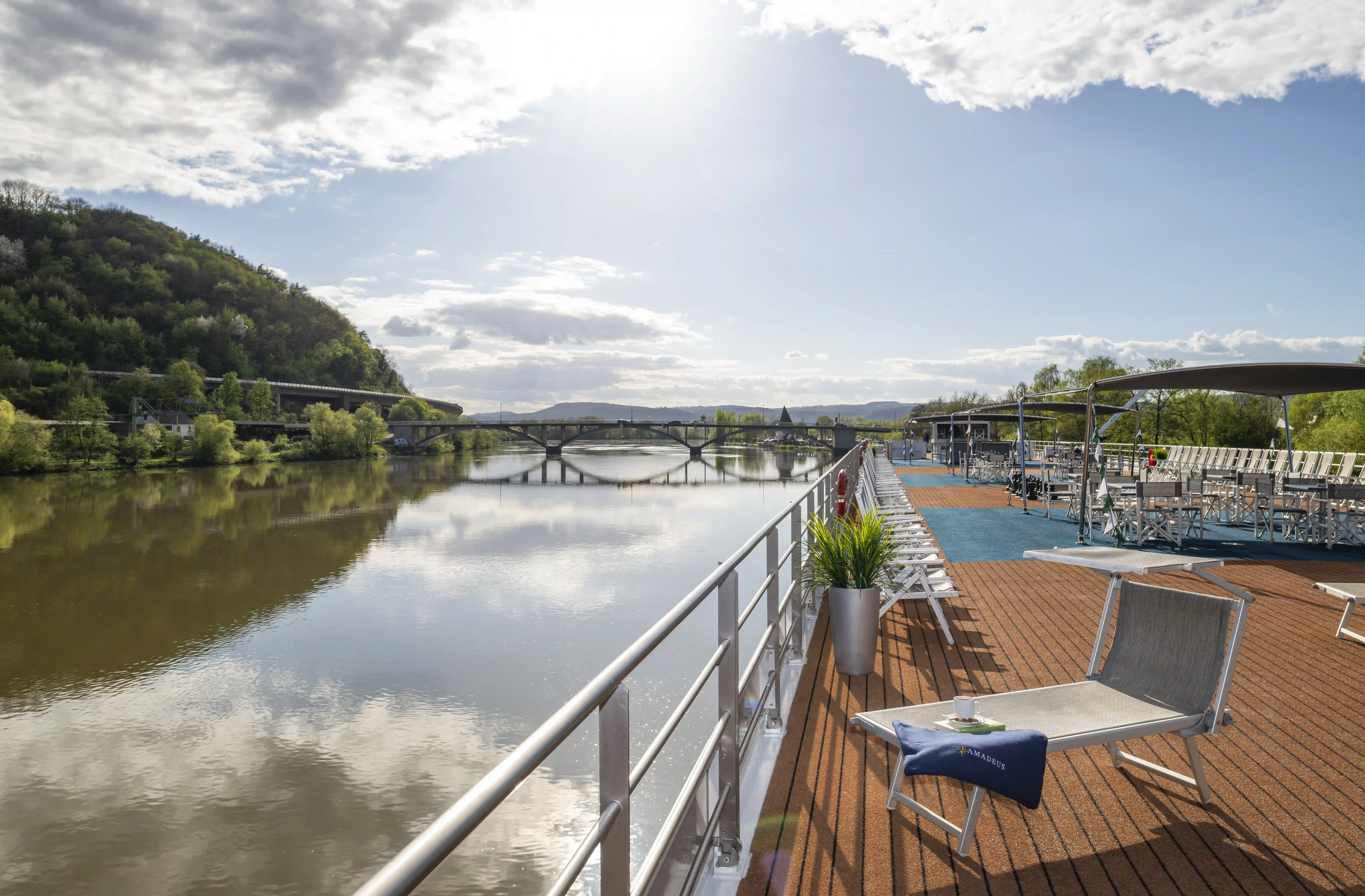 Sonnendeck eines Flussschiffes mit Liegestühlen, Sitzbereichen und Blick auf den ruhigen Fluss und eine Brücke