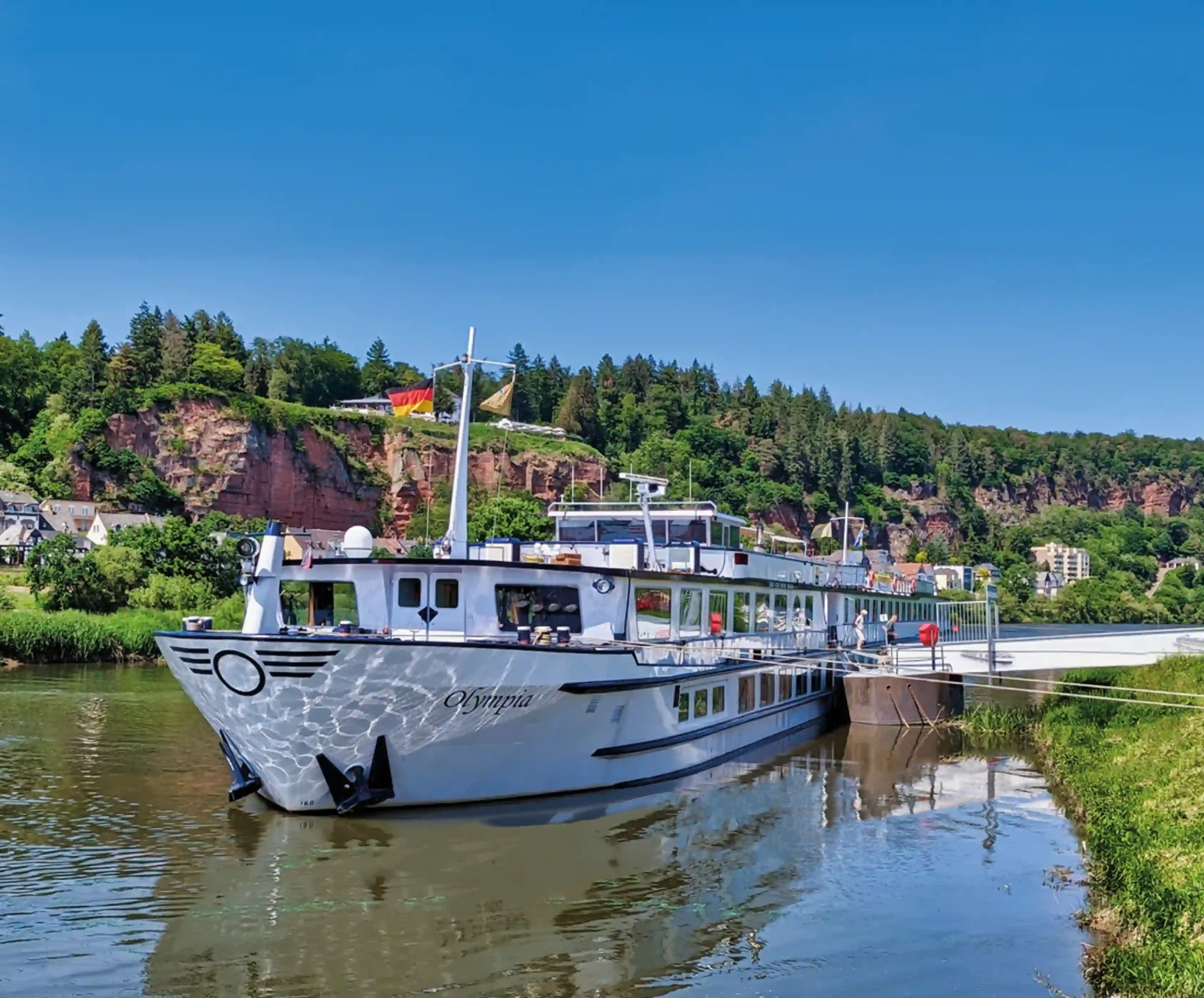Außenansicht des Flusskreuzfahrtschiffs „MS Olympia“, das an einem Anleger auf einem ruhigen Fluss liegt, umgeben von grünen Hügeln und Felsen bei sonnigem, blauem Himmel