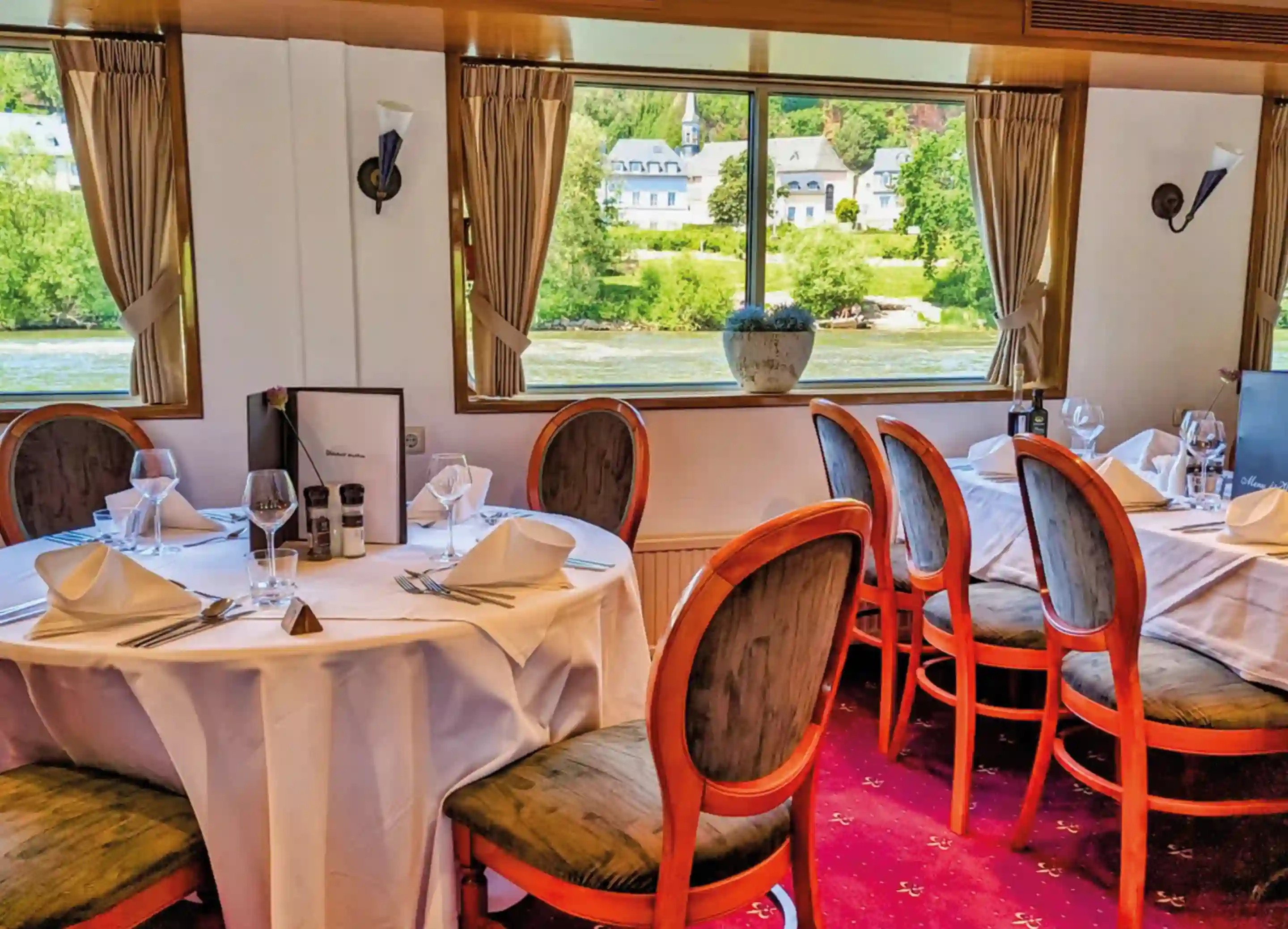 Elegant gedeckter Restaurantbereich an Bord der MS Olympia mit runden Tischen, weißen Tischdecken und großen Panoramafenstern mit Blick auf den Fluss und die Uferlandschaft