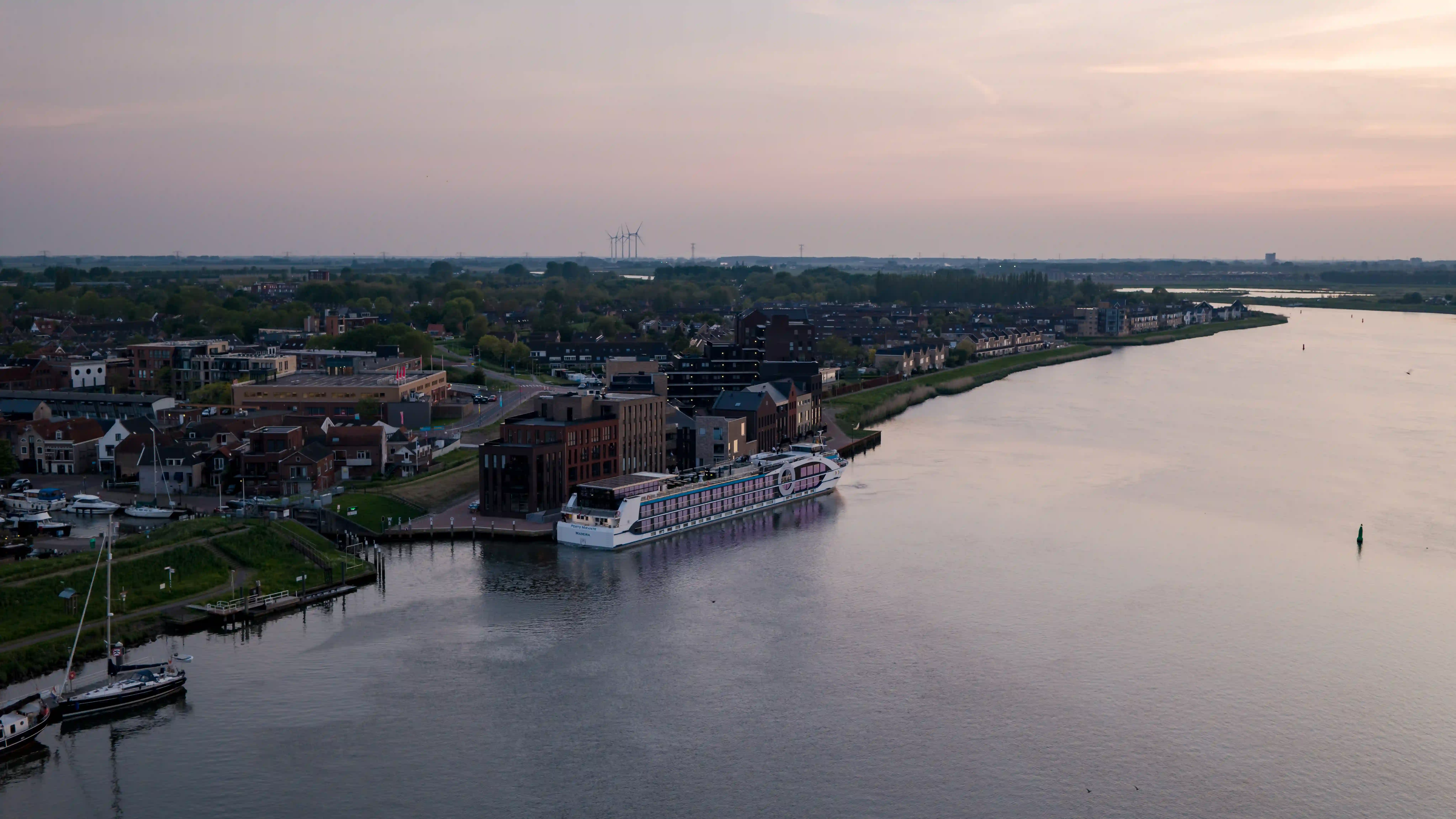 Flussschiff liegt bei Abenddaemmerung am Ufer eines breiten Flusses neben einer Stadt mit Wohnhäusern und Landschaft im Hintergrund.