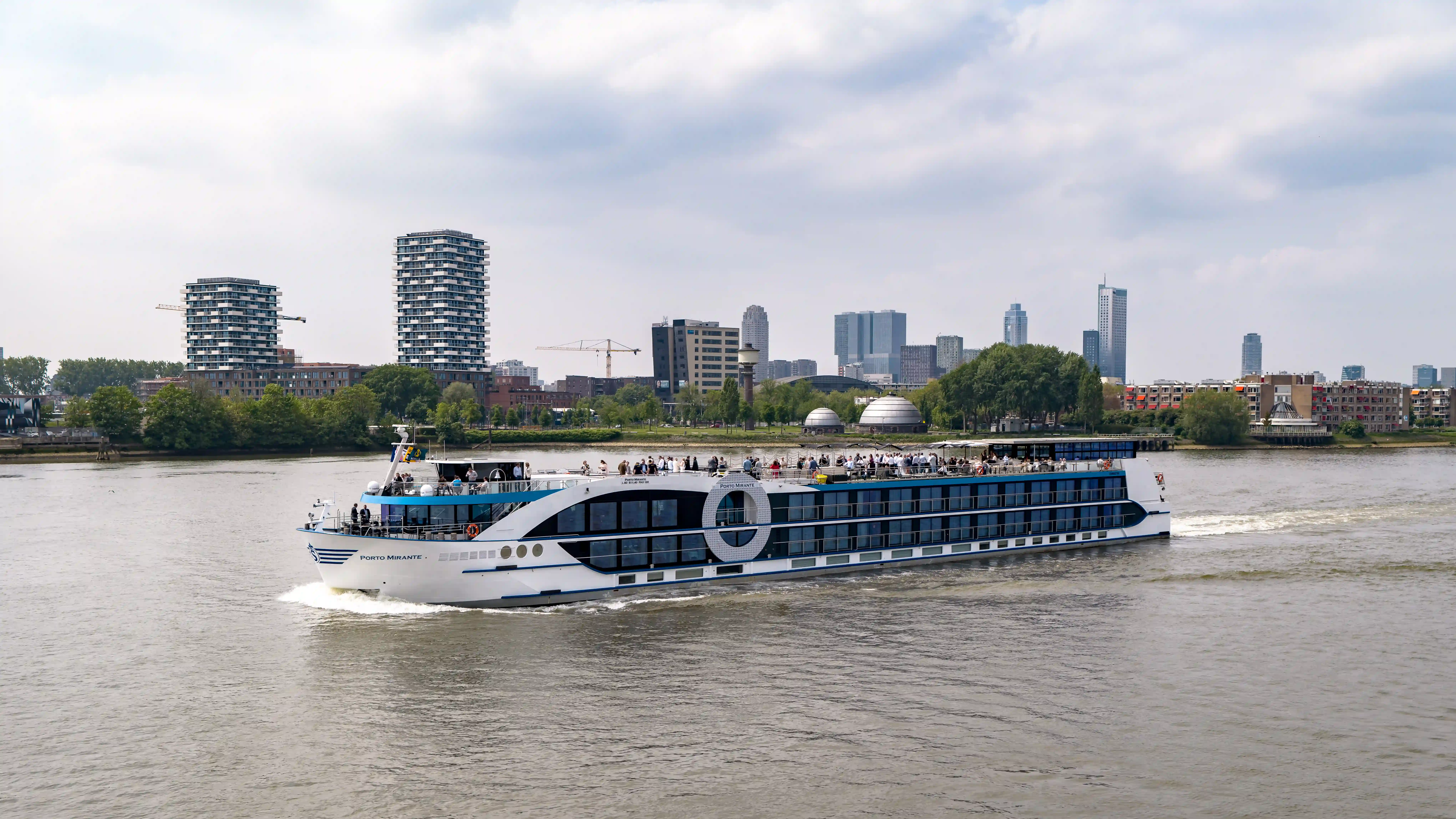 Modernes Flussschiff fährt auf breitem Fluss mit Stadtpanorama, Hochhäusern und Gruenanlagen im Hintergrund.