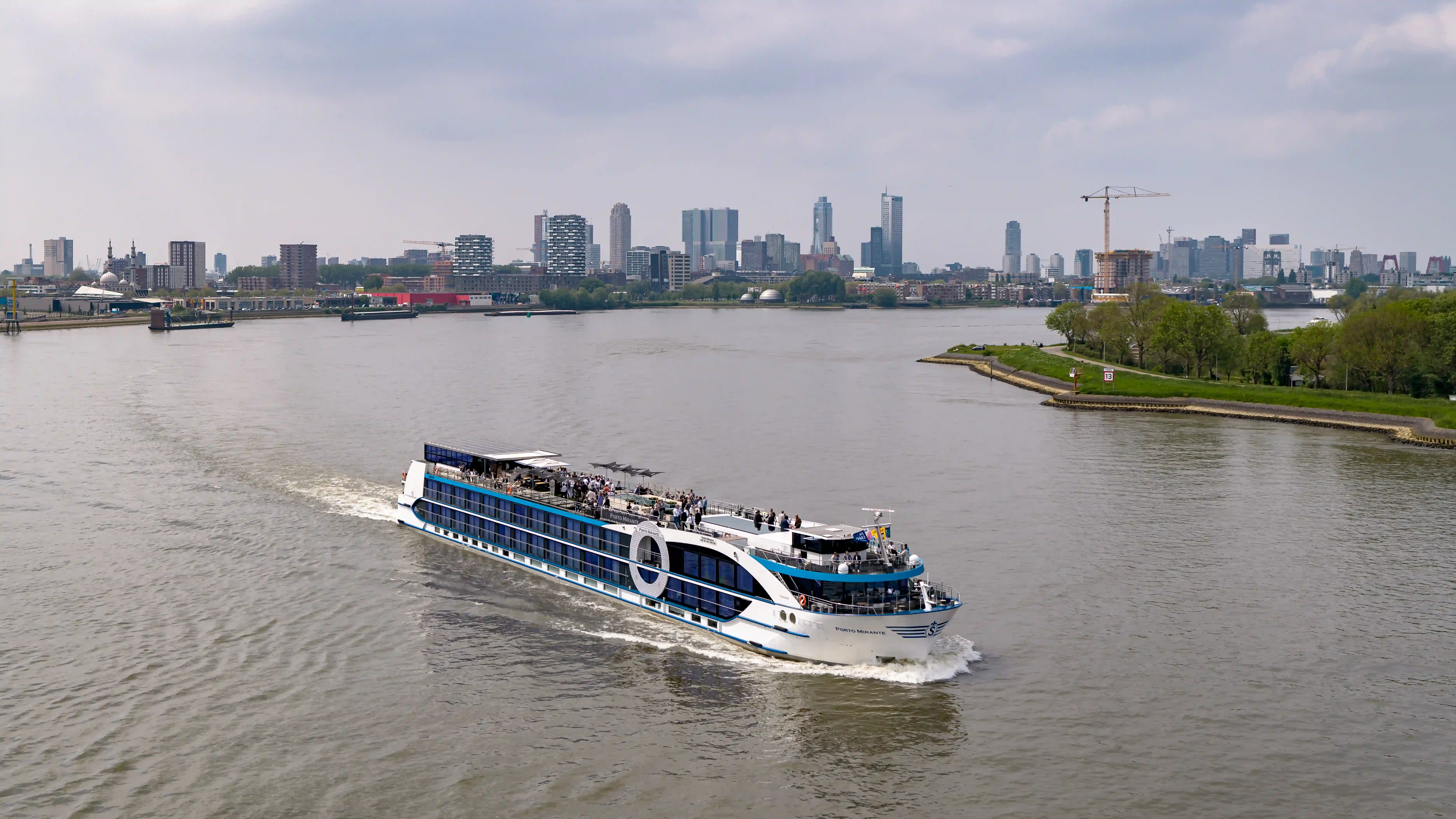 Flussschiff auf breitem Strom mit Blick auf Stadt, Industrieanlagen und gruene Uferlandschaft.