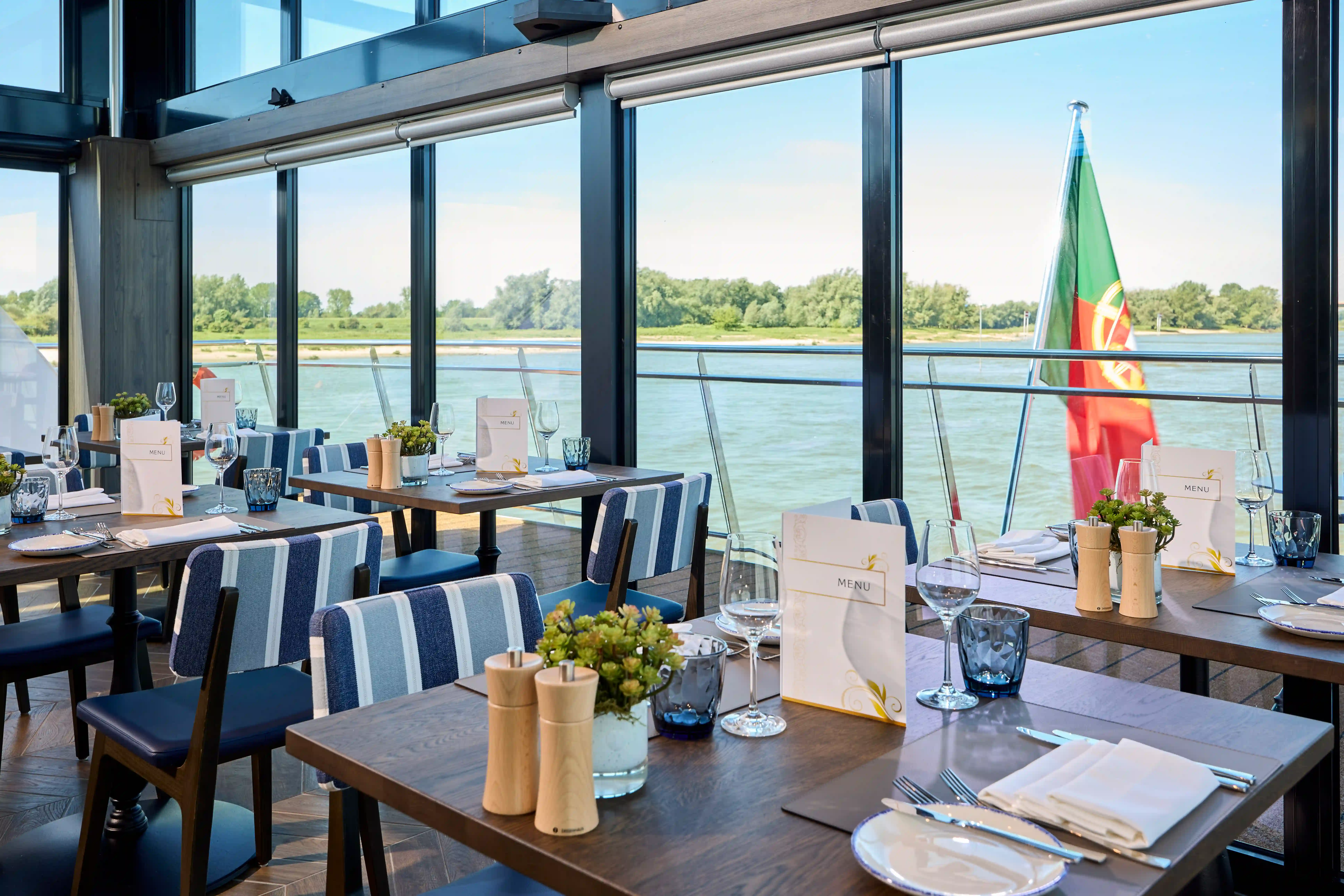 Bistrobereich mit gedeckten Tischen, großen Fenstern und Blick auf vorbeiziehende Flusslandschaft.
