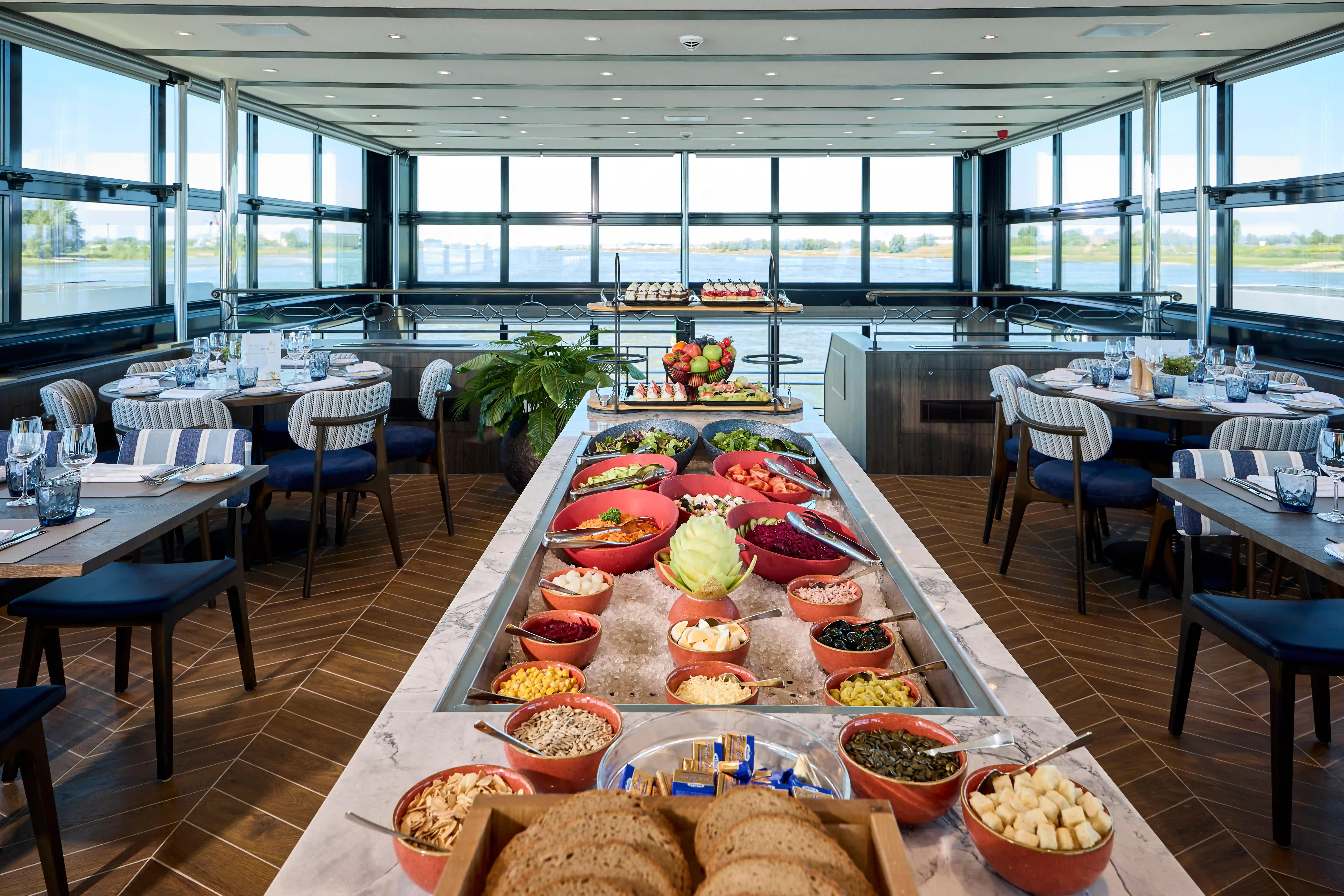 Bistro an Bord mit zentralem Buffet, Speisenanordnung und Sitzplätzen neben großen Fenstern.