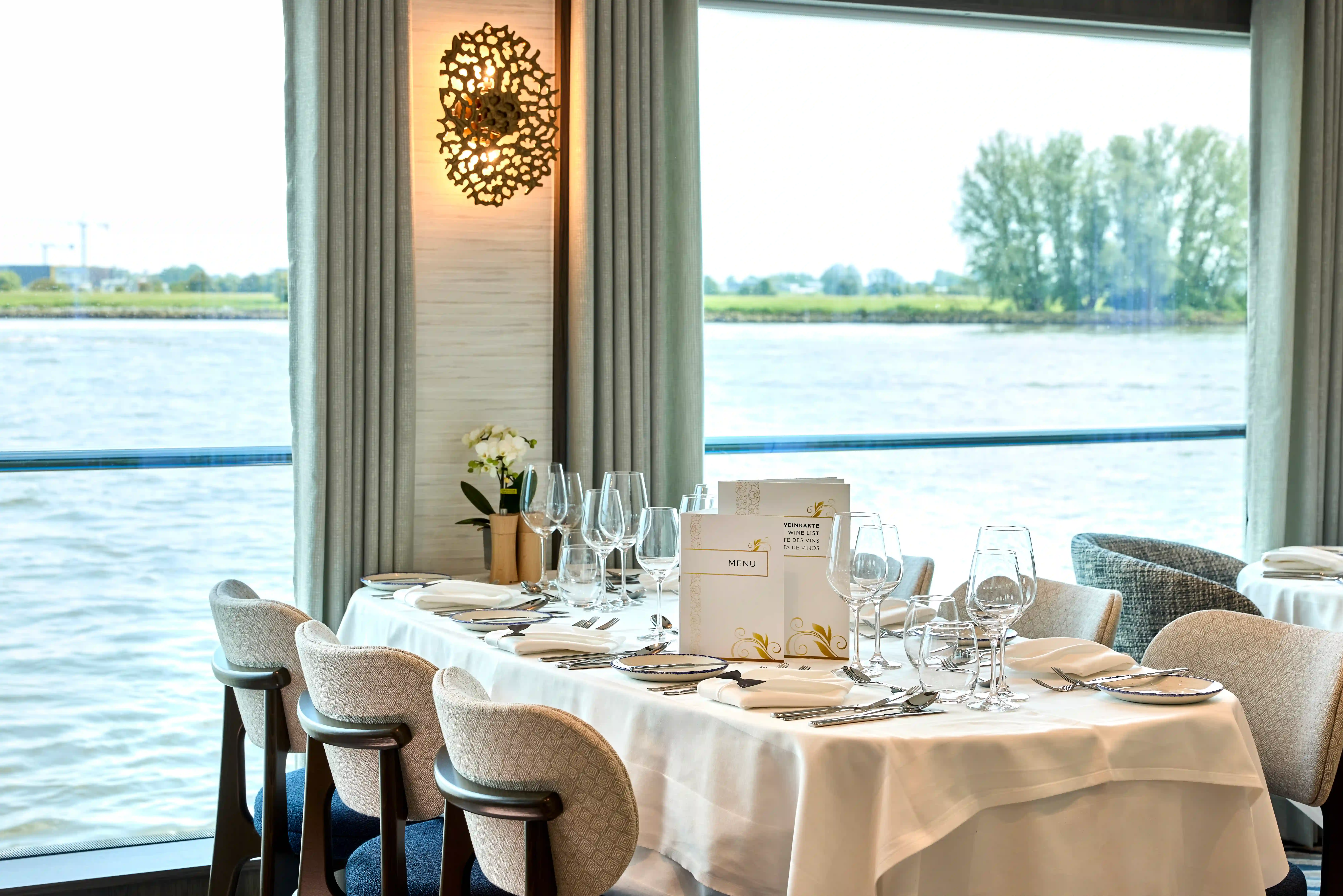 Gedeckter Restauranttisch mit Gläsern und Menükarte vor großen Fenstern mit Blick auf den Fluss und Uferlandschaft.