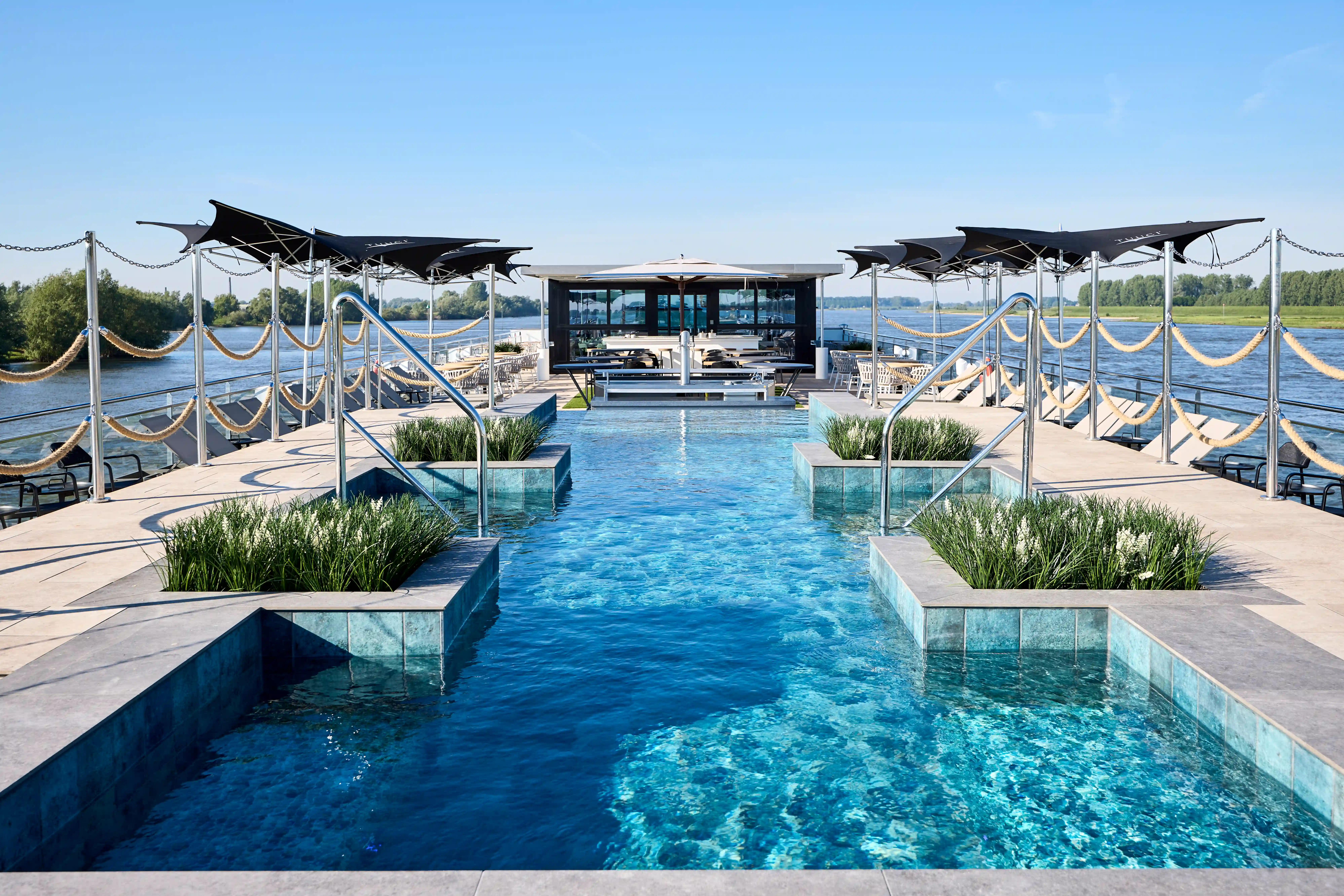 Sonnendeck eines Flussschiffes mit zentralem Pool, Liegeflächen, Sonnensegeln und Blick auf den Fluss.