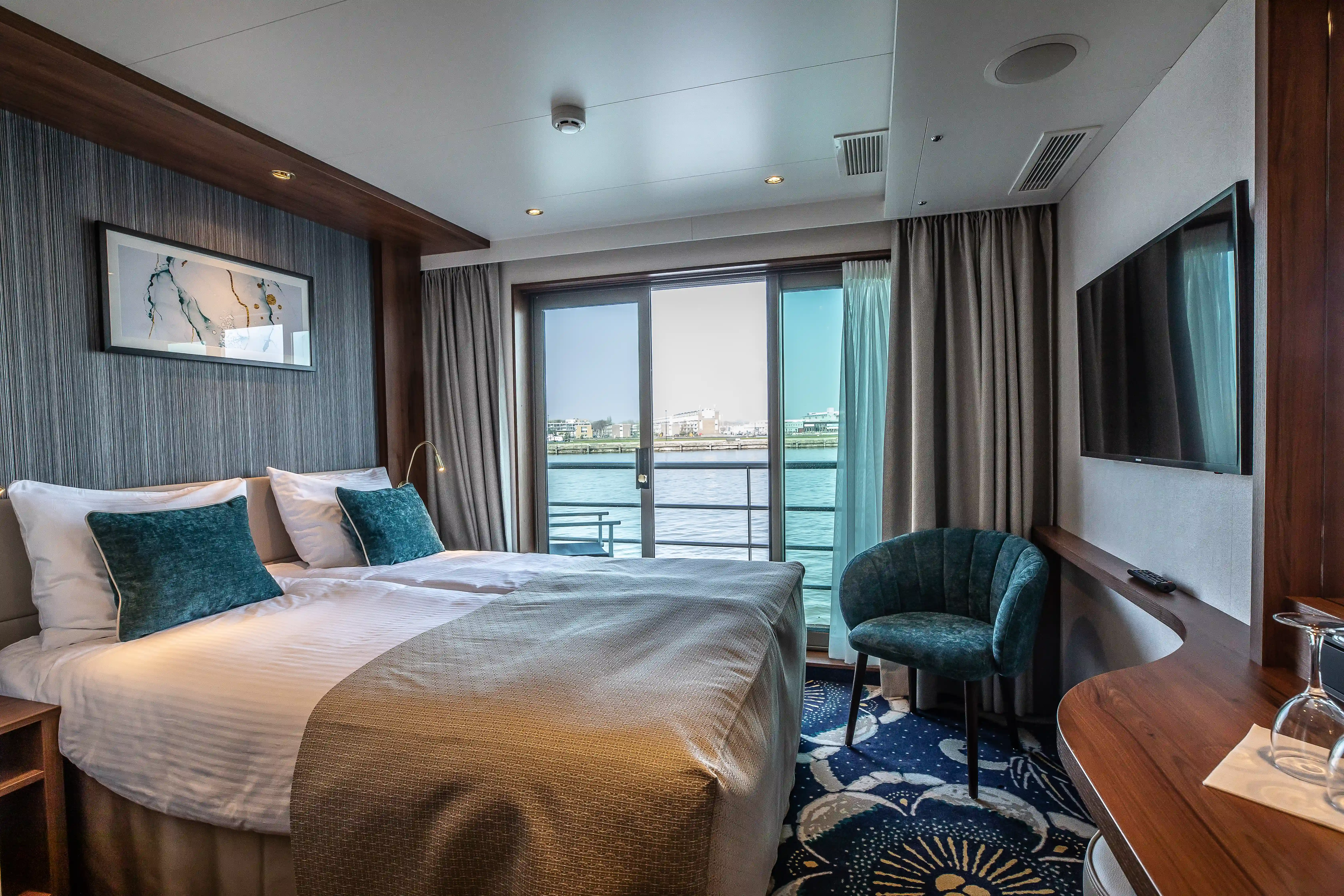 Moderne Schiffskabine mit Doppelbett, Sitzsessel, Schreibtisch und bodentiefem Fenster mit Blick auf den Fluss