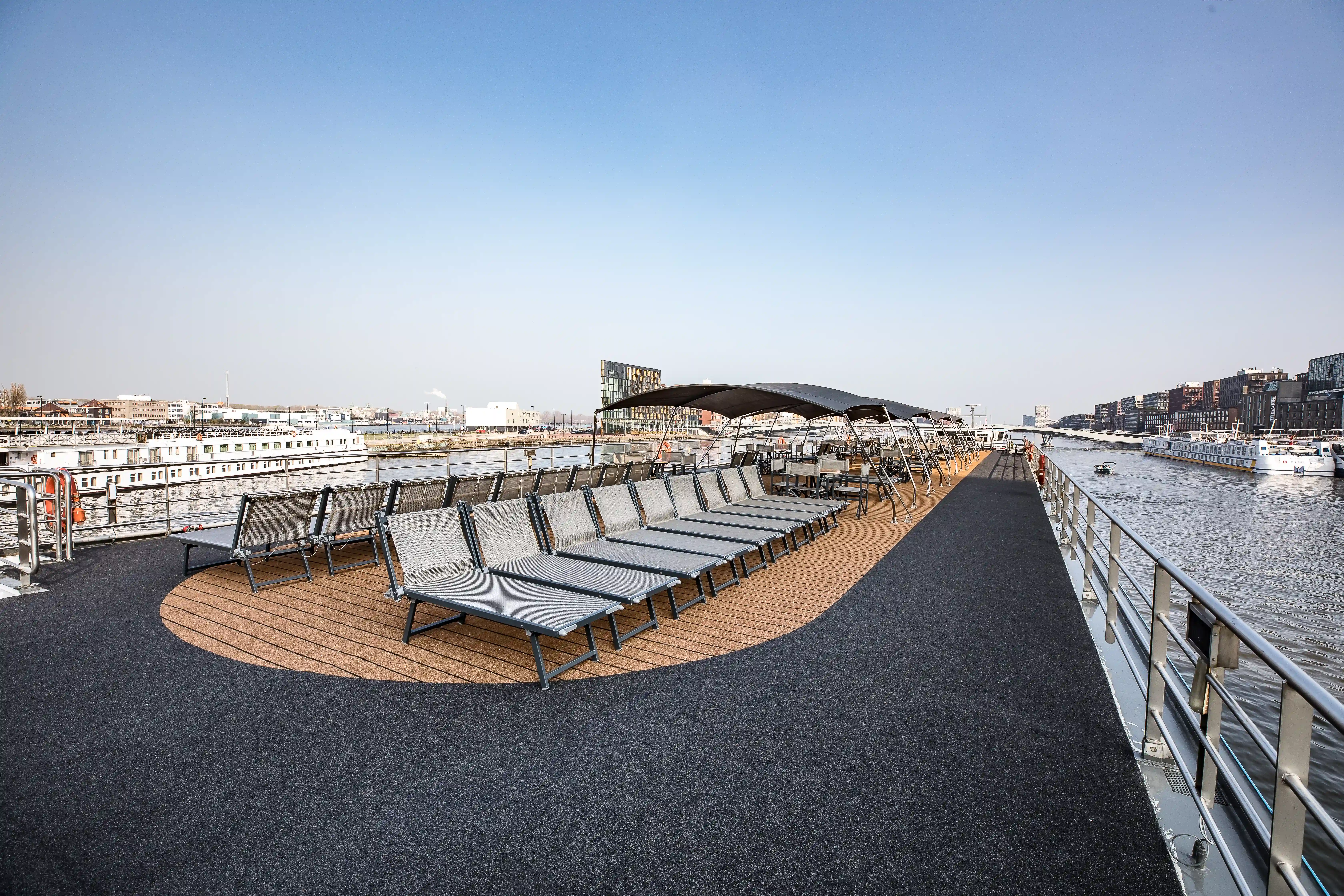 Sonnendeck mit Reihen von Sonnenliegen, schattenspendender Überdachung und Blick auf Hafen und Fluss
