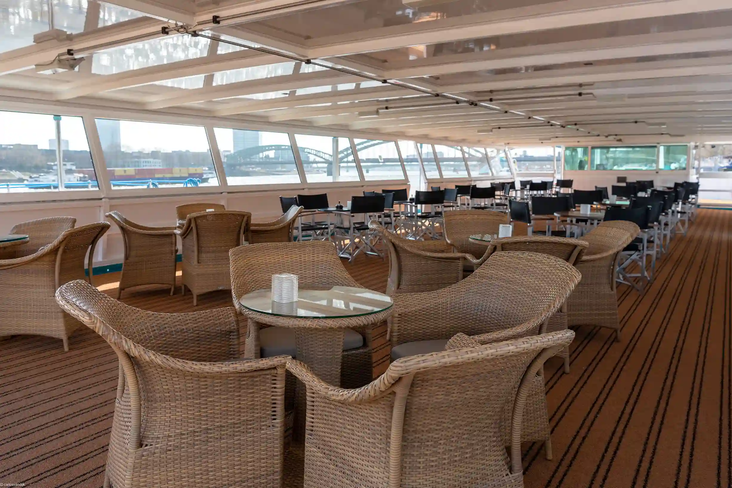 Überdachter Aussichtssalon auf dem Schiff mit hellem Holzboden, zahlreichen Sitzgruppen aus Korbsesseln und dunklen Regiestühlen an den Panoramafenstern.