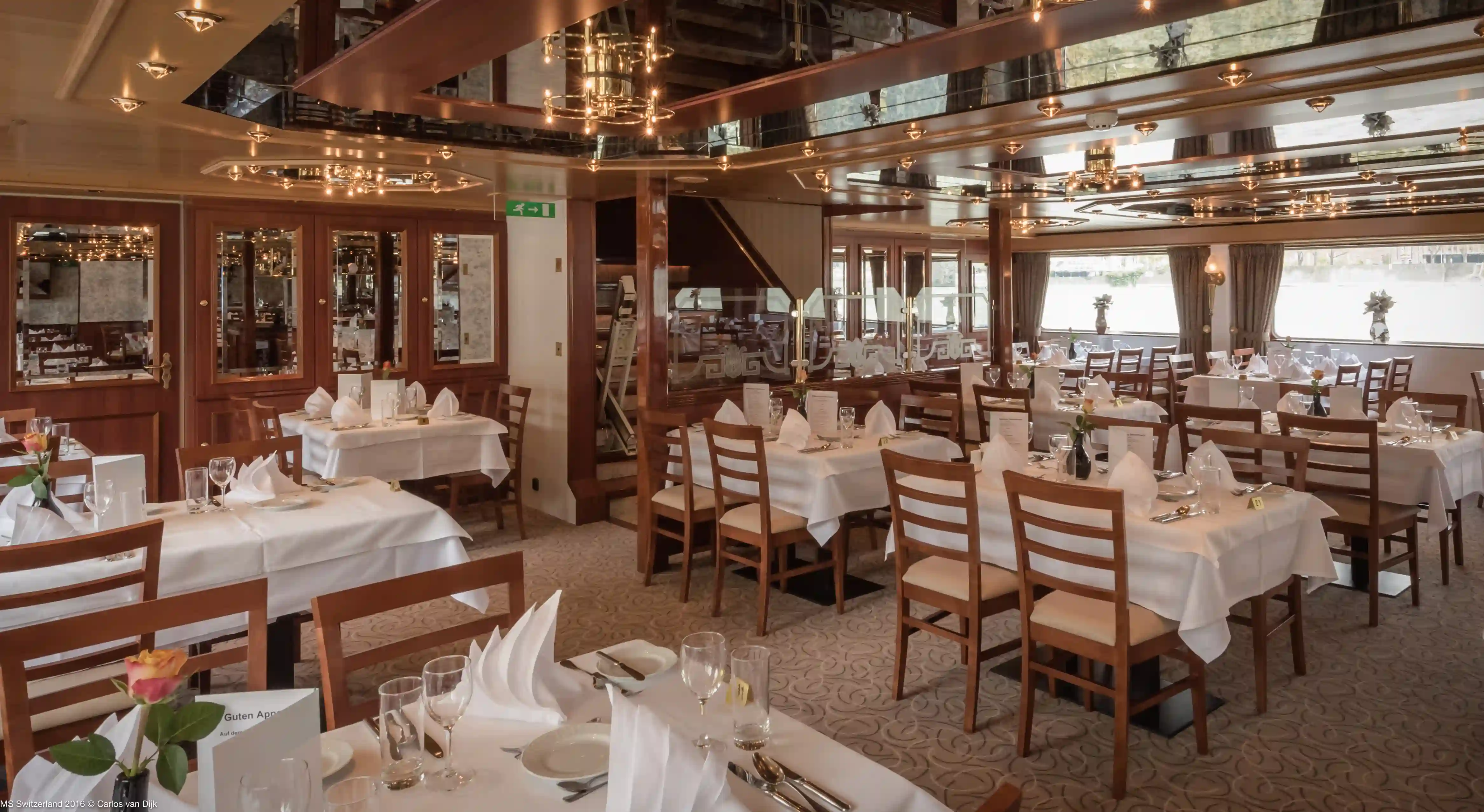 Elegant gedeckte Tische im Restaurant der MS Switzerland, getrennt durch eine kunstvolle Glaswand vor dem Treppenaufgang unter einer Spiegeldecke.