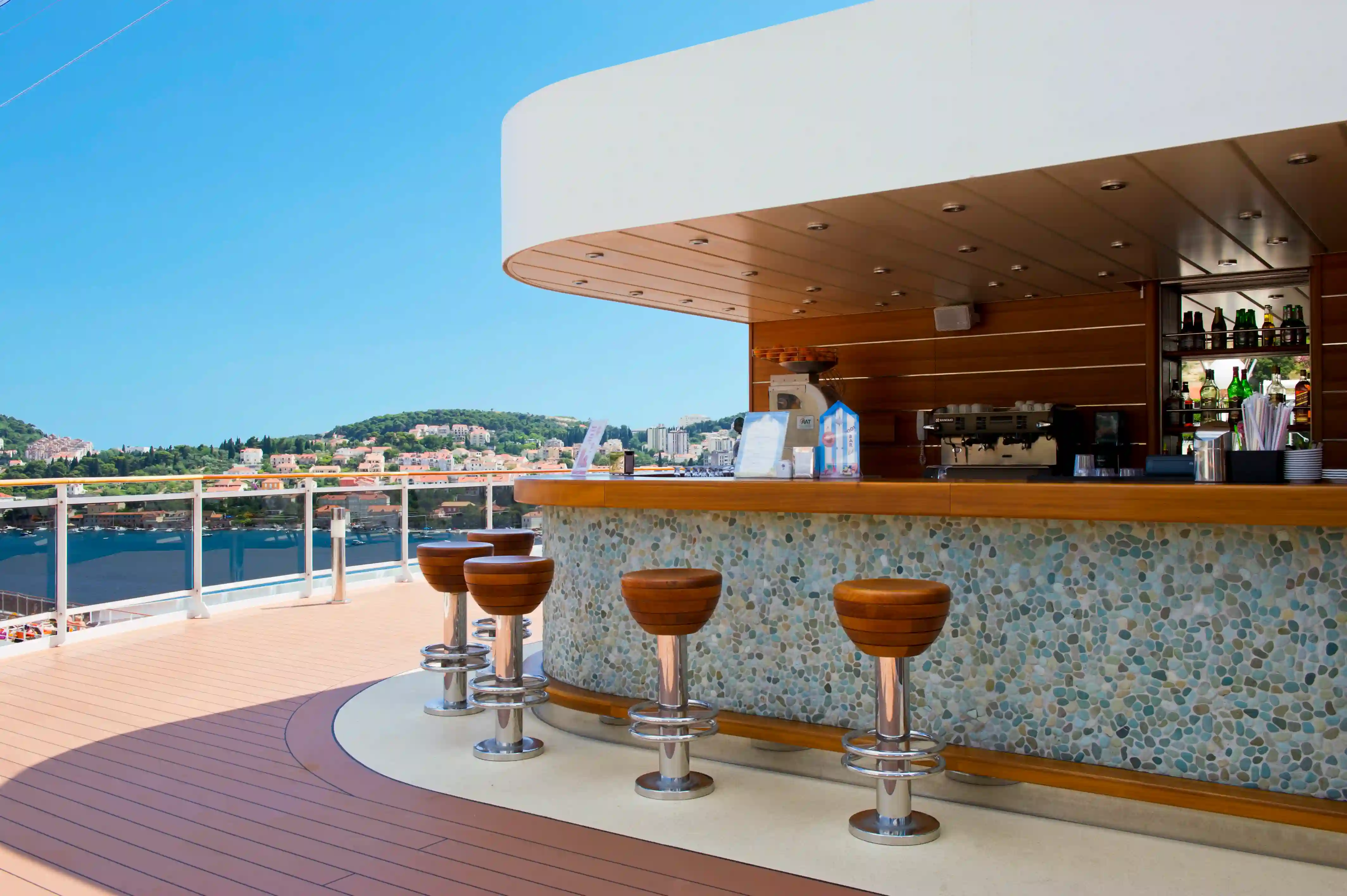 Aussenbar auf der MSC Magnifica mit Bartheke, Barhockern und Blick auf Hafen und Kuestenlandschaft.