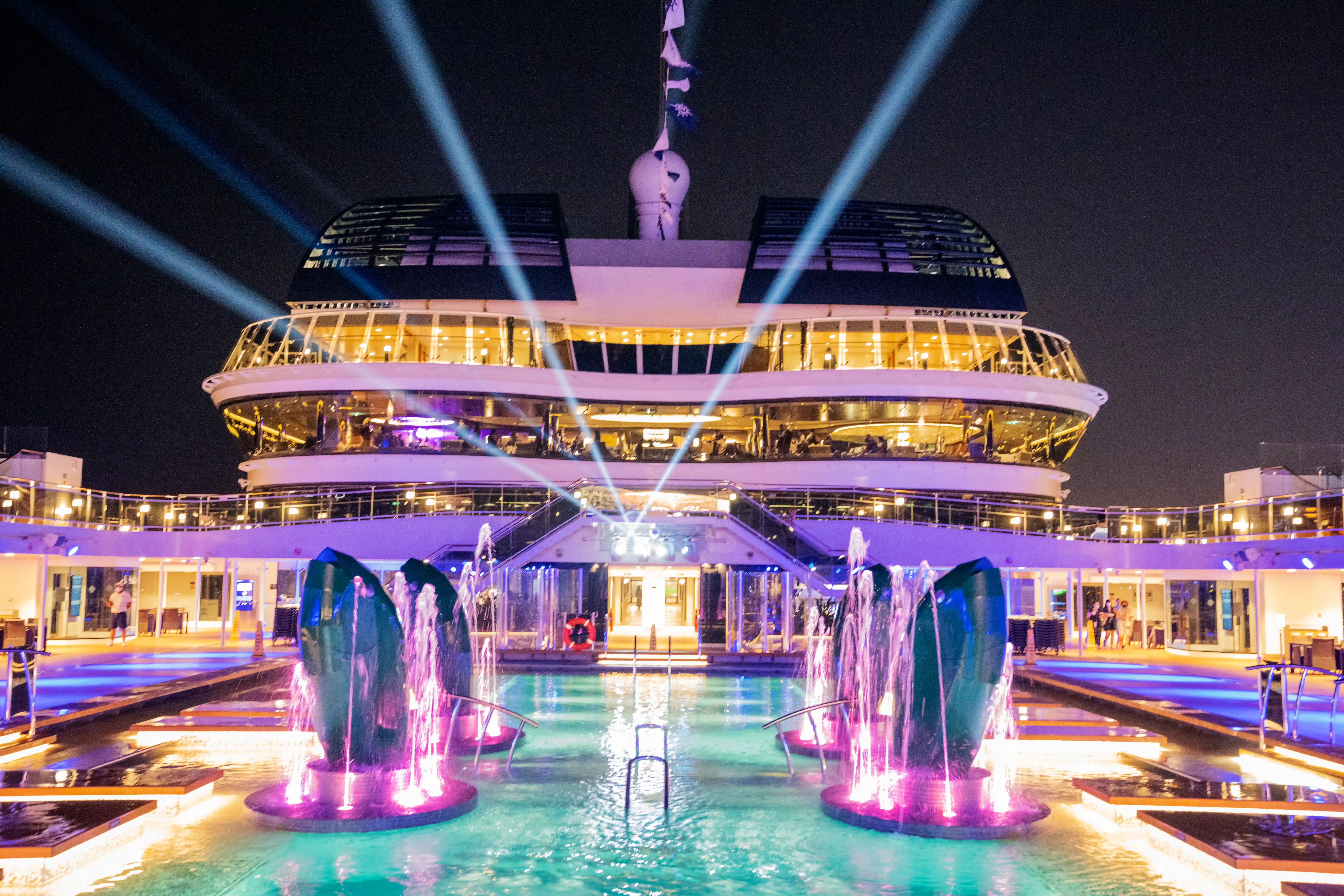 Aussenpool auf der MSC Virtuosa bei Nacht mit beleuchteten Wasserfontaenen und Deckbeleuchtung
