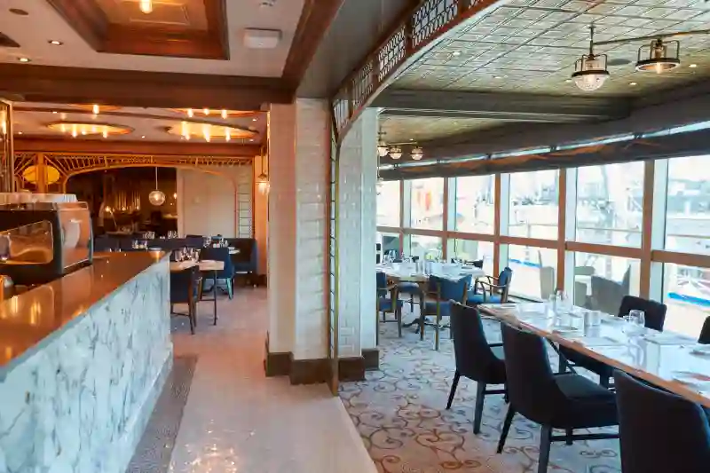 Ein elegant eingerichtetes Schiffsrestaurant mit gemustertem Teppich, dunklen Stühlen, einer edlen Marmortheke und großen Fensterfronten mit Blick nach draußen.